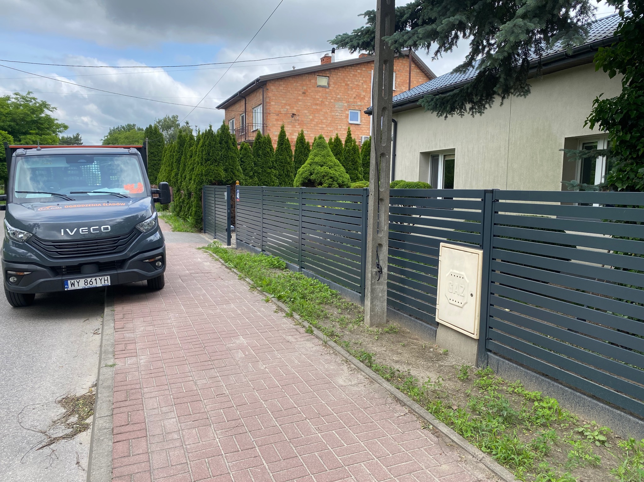 Nowoczesne, poziome ogrodzenie panelowe w kolorze antracytowym, widoczne z perspektywy chodnika. Na pierwszym planie samochód dostawczy firmy.