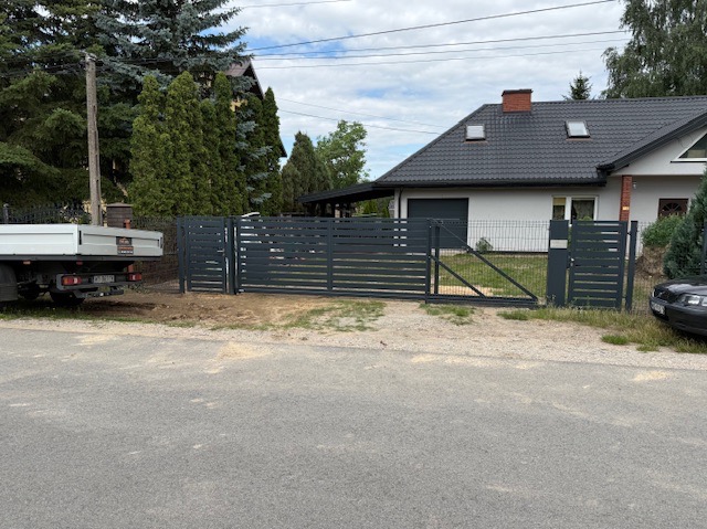 Nowoczesna, pozioma brama przesuwna i furtka w kolorze antracytowym, z elementami siatki, na tle domu z ciemną dachówką i trawnika. Widoczny fragment samochodu i auto dostawcze.