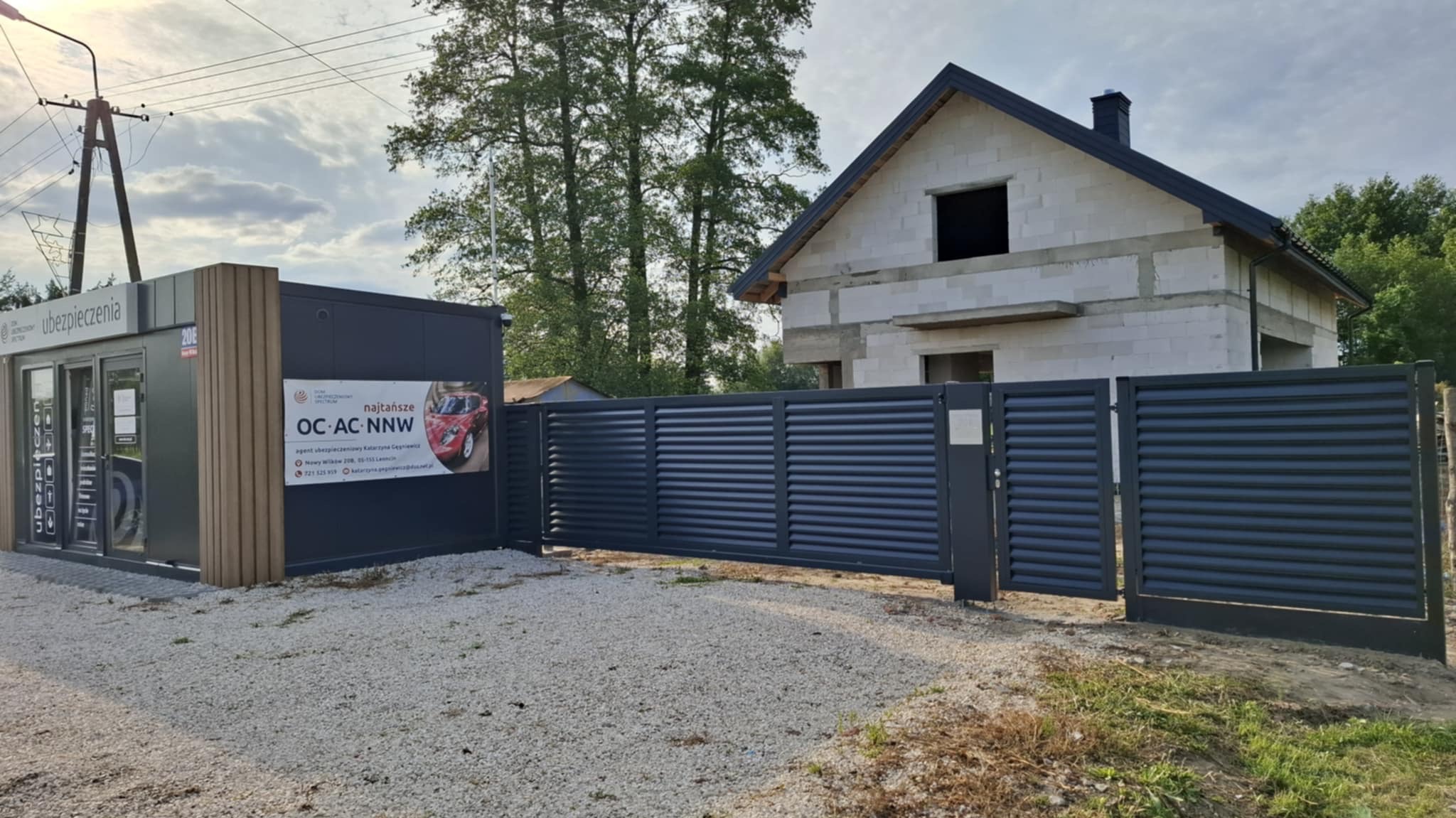 Nowoczesna, przesuwna brama wjazdowa z metalowymi panelami w kolorze grafitowym, na tle budynku w stanie surowym i budki ubezpieczeń.