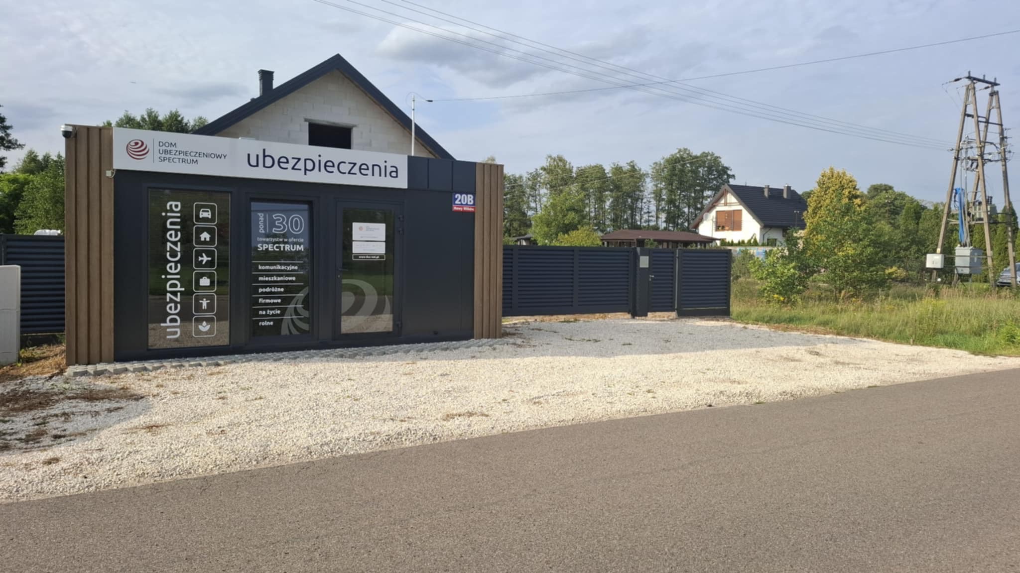Nowoczesne biuro ubezpieczeń z szarą, poziomą bramą w tle, zlokalizowane przy drodze, otoczone żwirowym podłożem. Widoczny budynek w budowie.