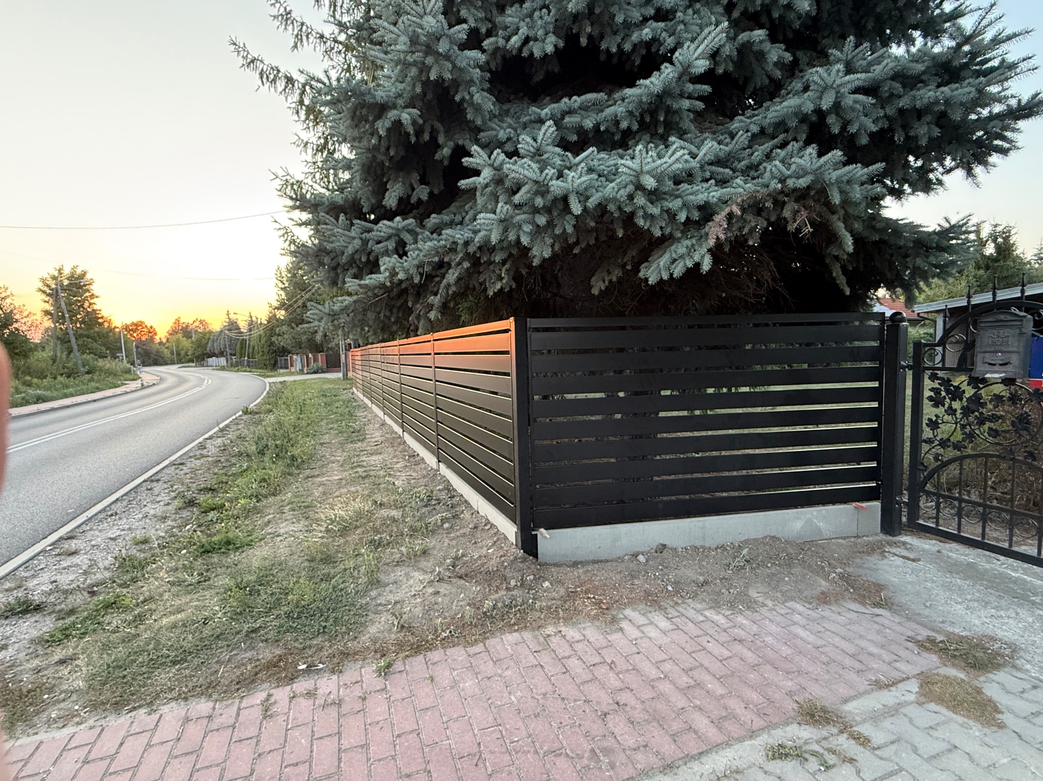 Nowoczesne, poziome ogrodzenie panelowe w kolorze antracytowym, z bramą w tle, otoczone zielenią i kostką brukową. Ujęcie z perspektywy ulicy, widoczne drzewo.