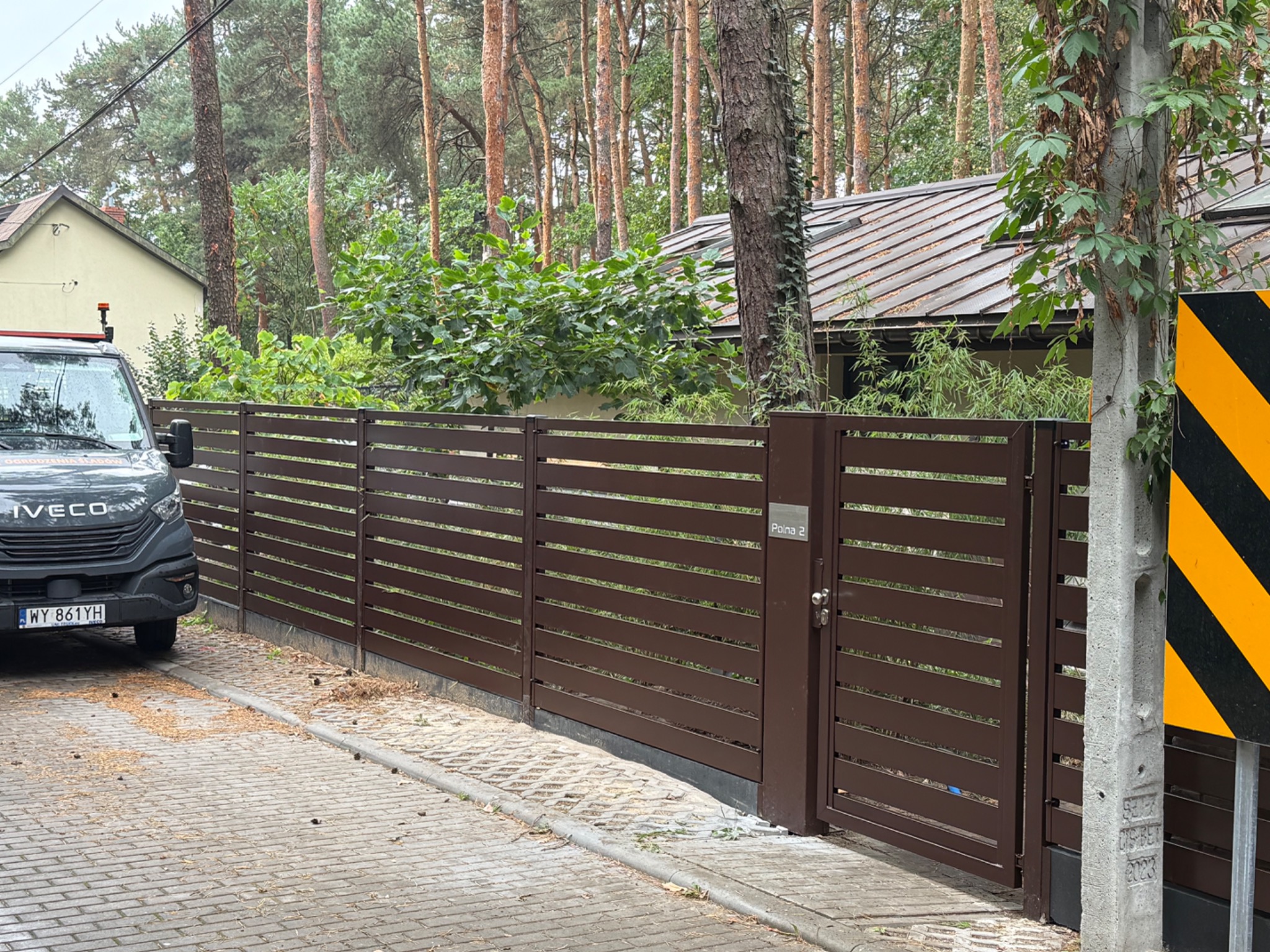 Nowoczesne, poziome ogrodzenie panelowe w kolorze brązowym z furtką i numerem domu 'Polna 2', na tle drzew i częściowo widocznego busa.
