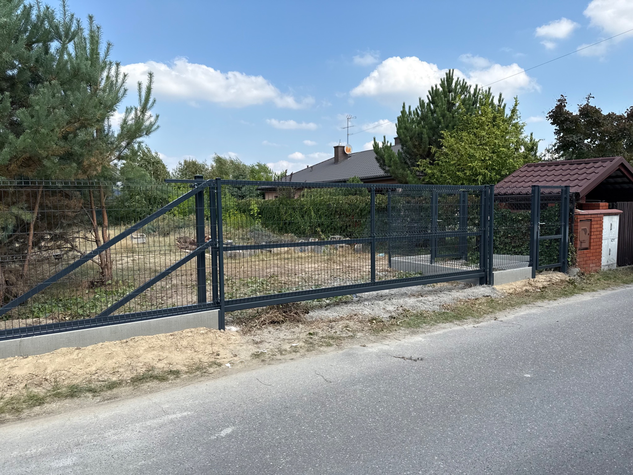 Nowoczesna, przesuwna brama panelowa w kolorze grafitowym, zainstalowana na betonowej podmurówce. Widok z ulicy, w tle dom i zieleń.