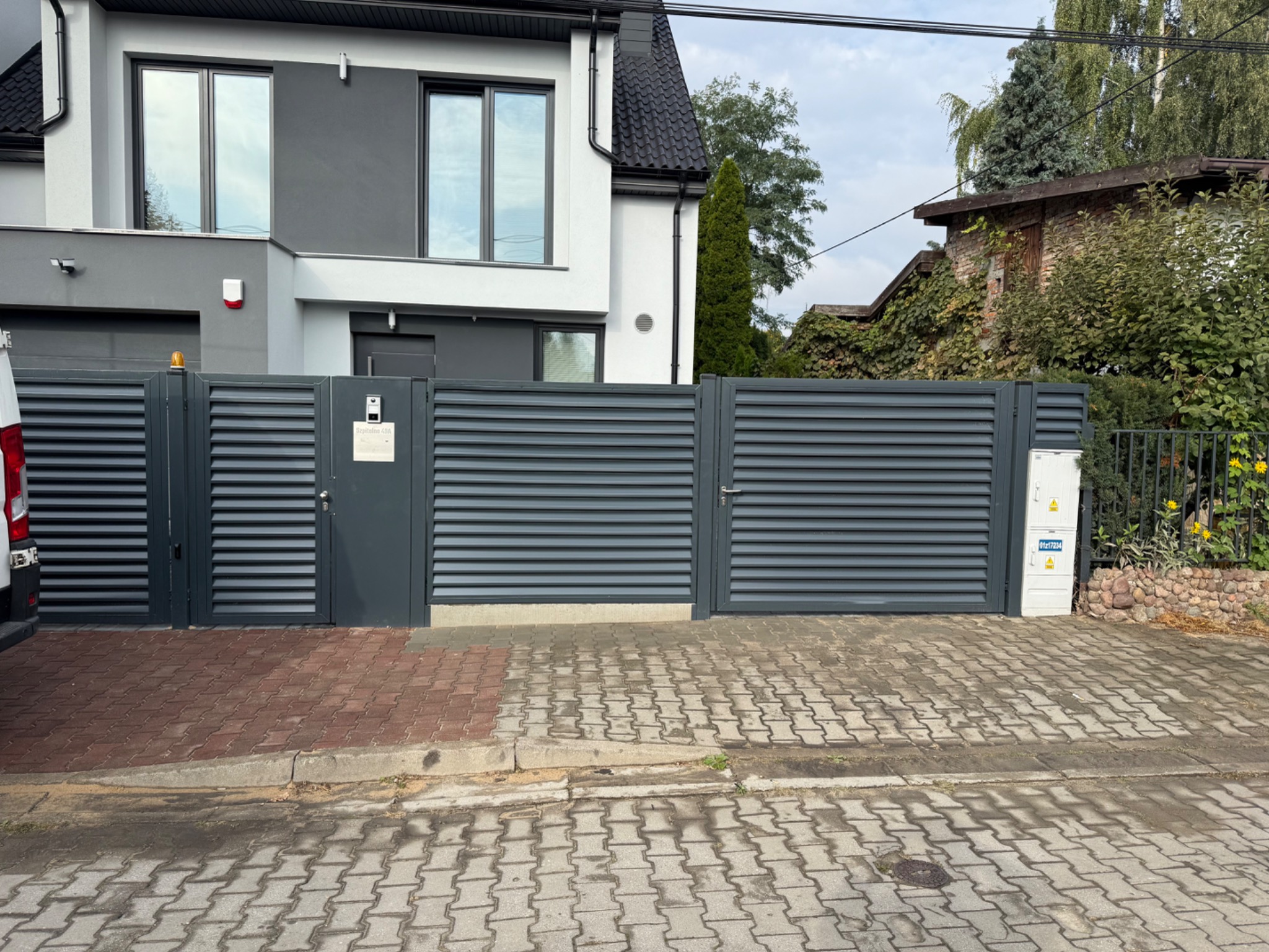 Nowoczesna, pozioma brama lamelowa w kolorze antracytu, z furtką i skrzynką elektryczną, na tle domu z szarą elewacją i kostki brukowej.