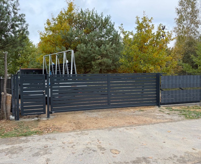 Nowoczesna, przesuwna brama wjazdowa w kolorze grafitowym z poziomymi panelami, furtka po lewej stronie. W tle drzewa i konstrukcja metalowa na pace samochodu.