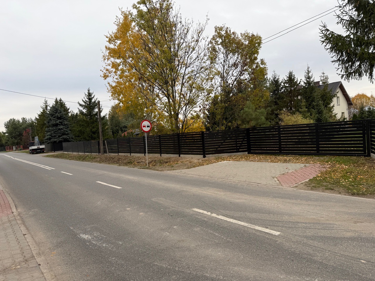 Długa, pozioma, czarna palisada. Nowoczesne ogrodzenie z poprzecznymi szczeblami, widok z ulicy, jesienne drzewa w tle, znak drogowy.