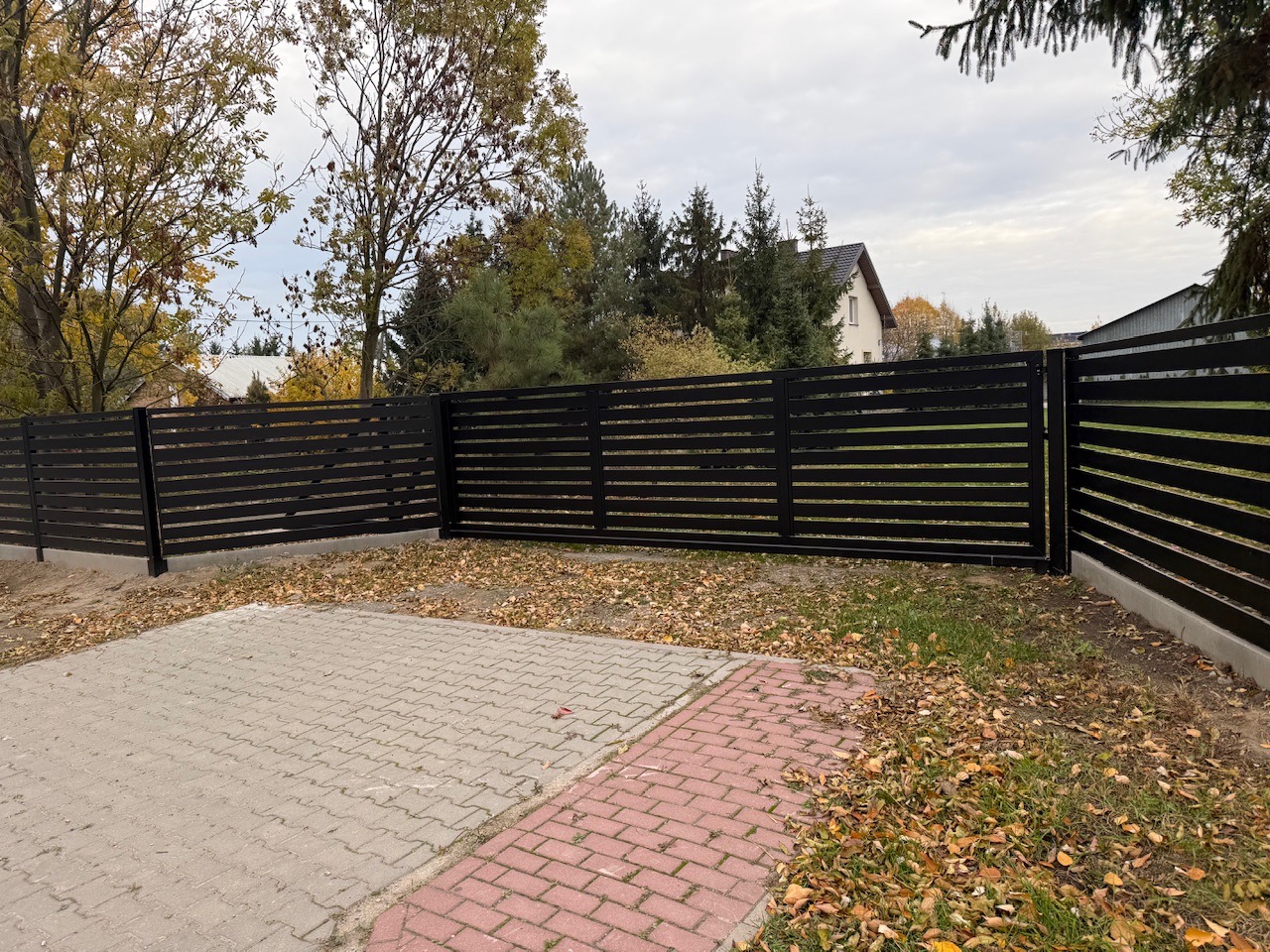 Nowoczesne, czarne ogrodzenie posesji z poziomych paneli, widoczne z zewnątrz na tle jesiennego krajobrazu i kostki brukowej. Minimalistyczny design.