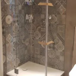 Nowoczesna kabina prysznicowa z deszczownicą, wykończona płytkami w geometryczne wzory. Widoczne półki z kosmetykami i fragment toalety. Elementy zabezpieczone taśmą malarską.