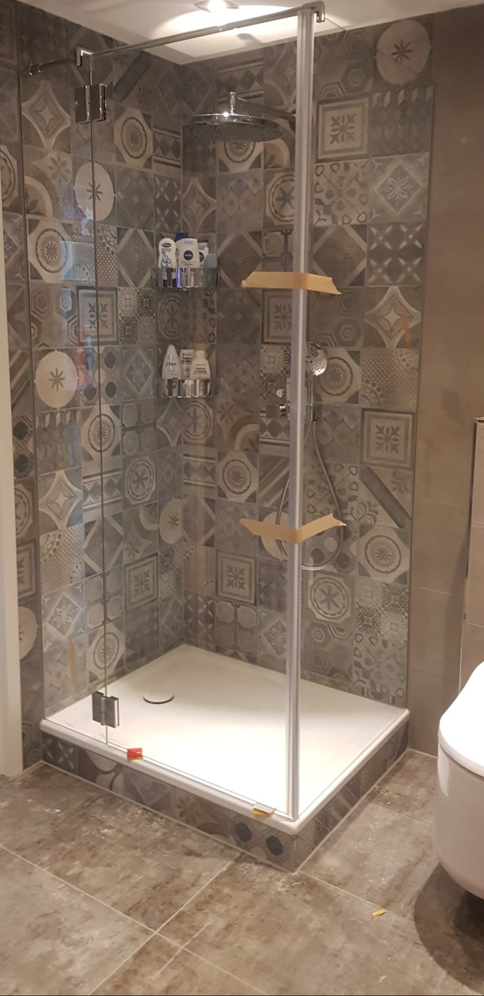 Nowoczesna kabina prysznicowa z deszczownicą, wykończona płytkami w geometryczne wzory. Widoczne półki z kosmetykami i fragment toalety. Elementy zabezpieczone taśmą malarską.
