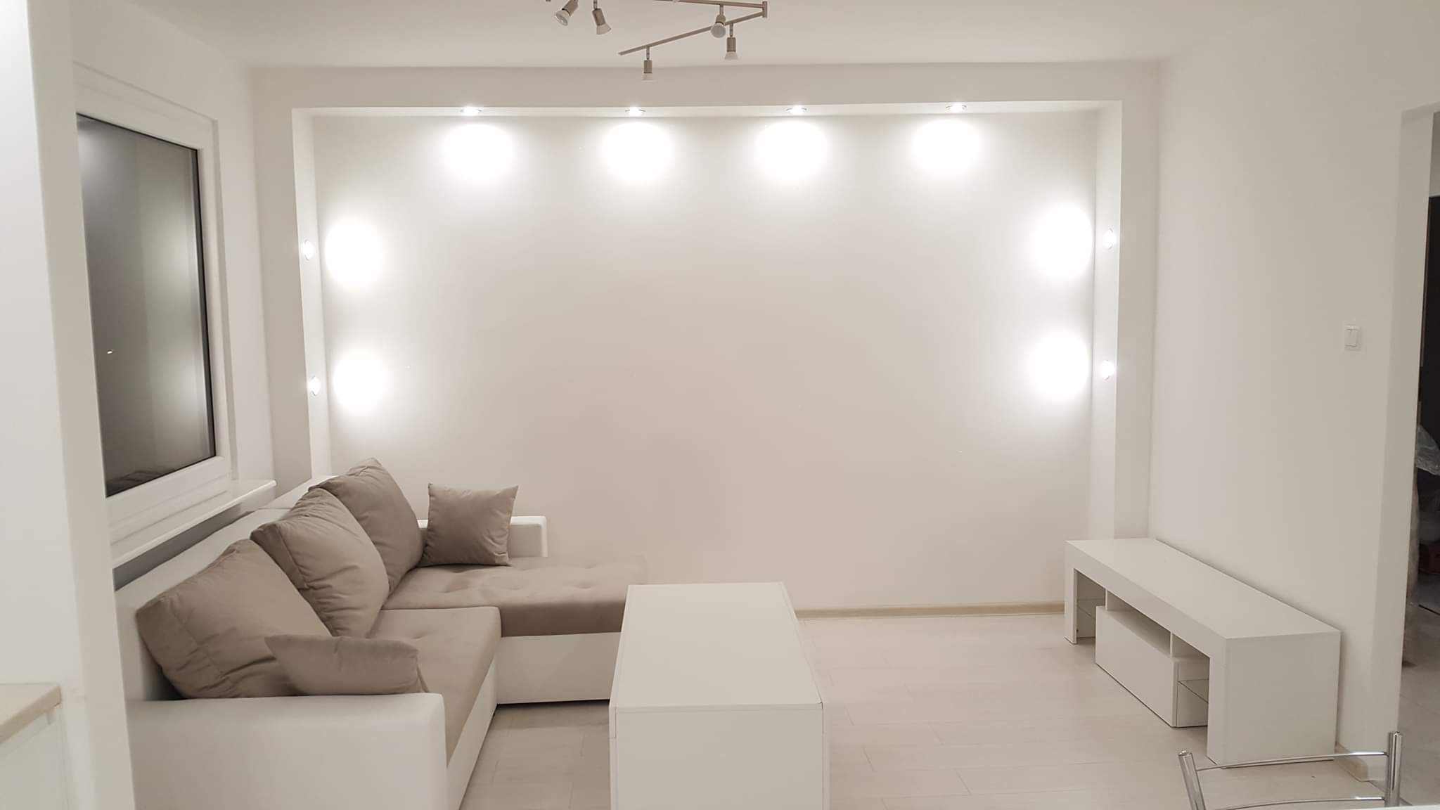 Jasne, minimalistyczne wnętrze po remoncie: białe ściany z wnęką oświetloną punktowymi lampami, narożna sofa w odcieniach beżu, biały stolik kawowy i szafka RTV, parkiet w jasnym odcieniu drewna.