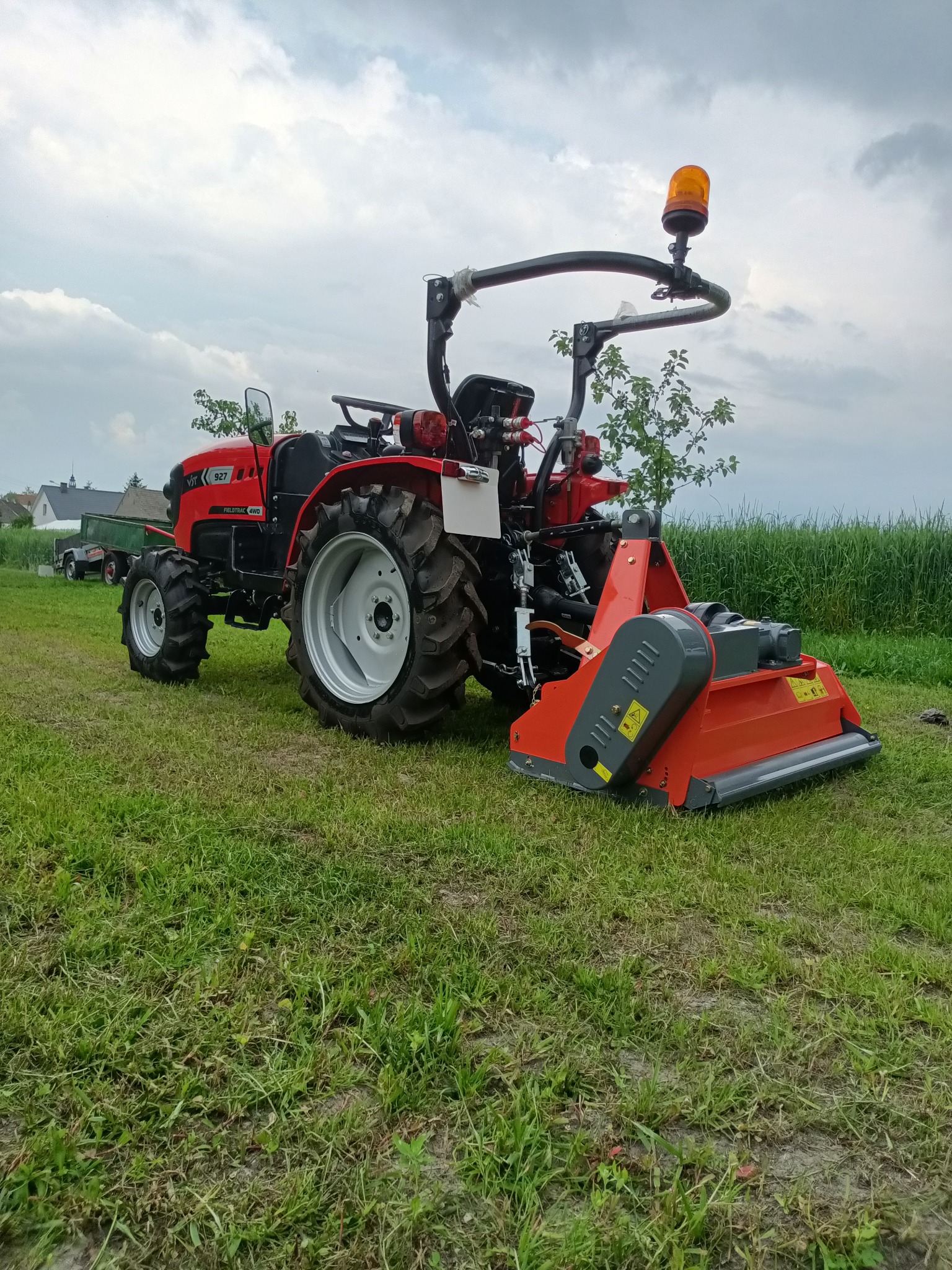 Czerwony traktor z kosiarką rotacyjną na zielonym trawniku, widoczne budynki w tle pod pochmurnym niebem.