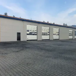 Szereg segmentowych bram wjazdowych do hali przemysłowej z szarymi panelami i białymi ramami, widok z zewnątrz na brukowany plac.