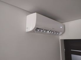 Biały klimatyzator ścienny zamontowany w rogu pokoju z jasnymi ścianami, widoczny fragment ciemnych drzwi po prawej stronie.