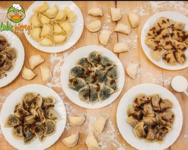 Widok z góry na drewniany stół z pięcioma talerzami pierogów o różnym nadzieniu i posypanych smażoną cebulką, otoczonych surowymi pierogami posypanymi mąką; na jednym z talerzy pierogi z kapustą...