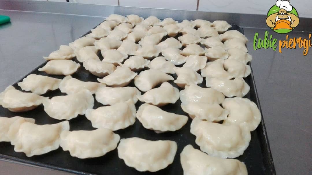 Blacha pełna świeżo ulepionych, błyszczących pierogów z nadzieniem, widoczne logo firmy 'lubię pierogi' w prawym górnym rogu.
