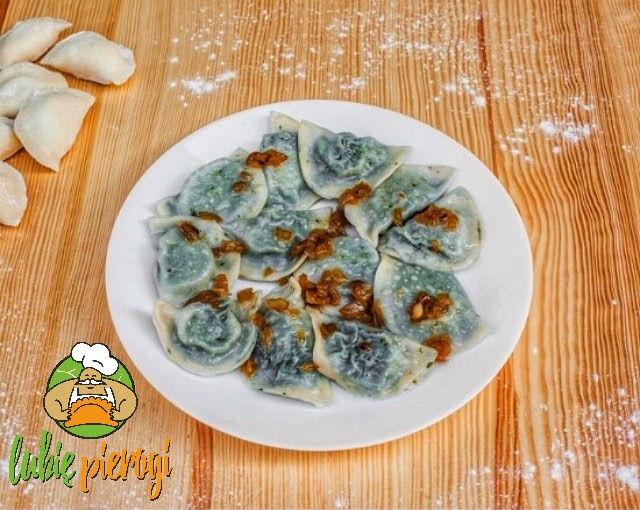 Porcja pierogów z farszem szpinakowym, polanych podsmażoną cebulką, ułożonych na białym talerzu, na drewnianym stole z posypką mąki. W lewym dolnym rogu logo firmy z napisem 'lubię pierogi'.