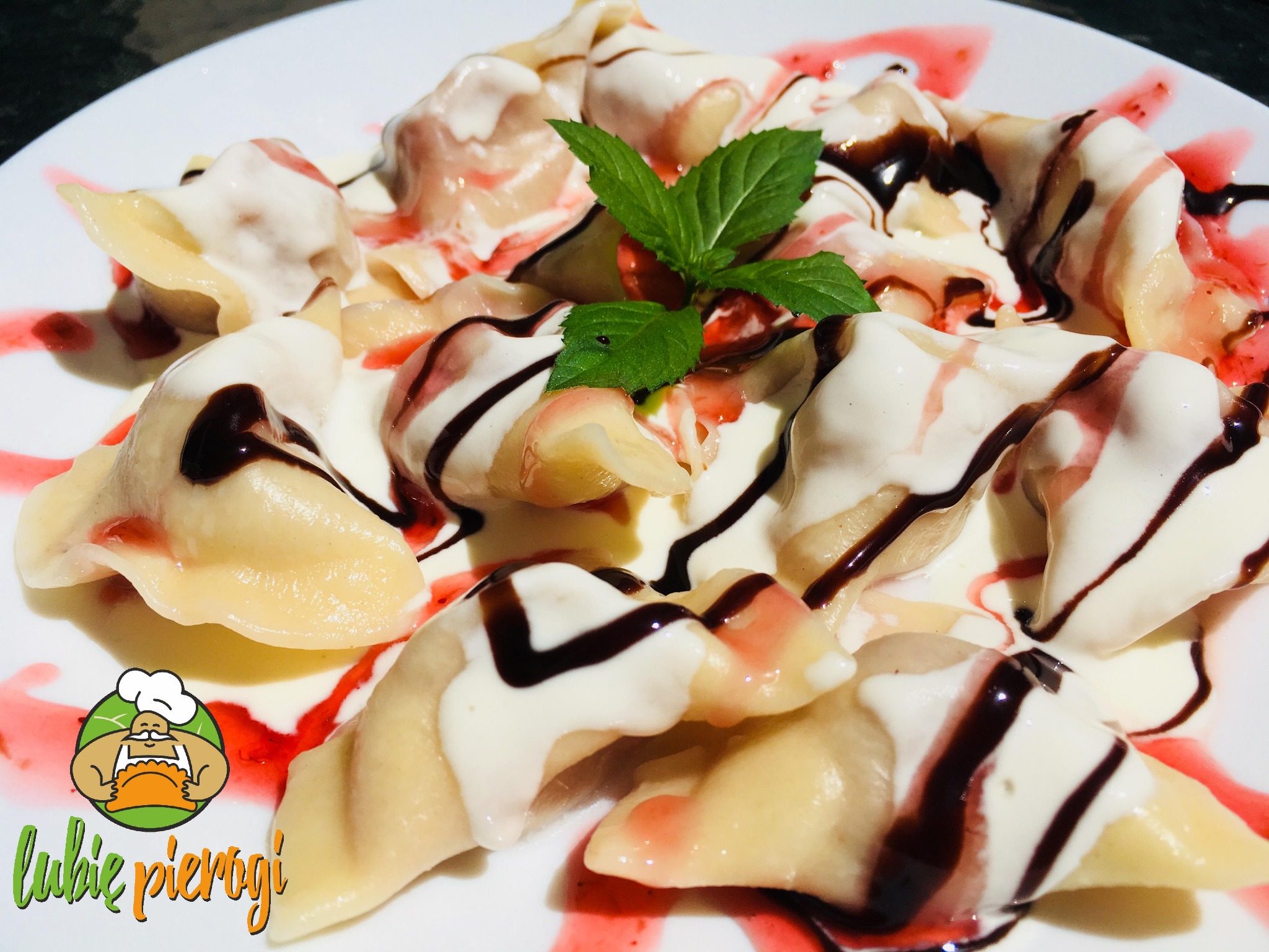 Porcja pierogów polanych śmietaną, sosem czekoladowym i syropem owocowym, udekorowana listkiem mięty na białym talerzu, z widocznym logo firmy 'lubie pierogi'.