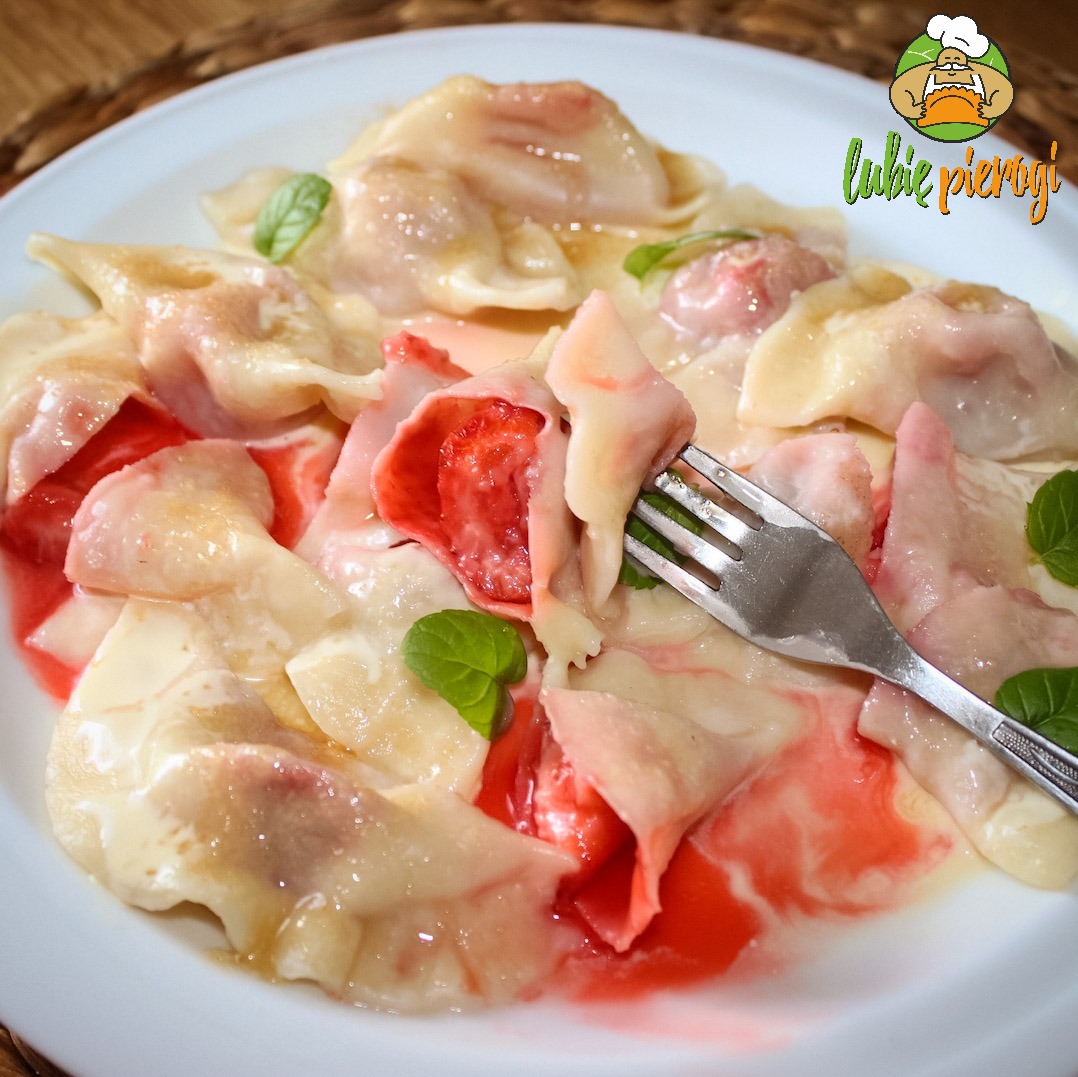 Porcja pierogów z truskawkami na białym talerzu, jeden pieróg nadziany na widelec, dekoracja z listków mięty, logo firmy w prawym górnym rogu.