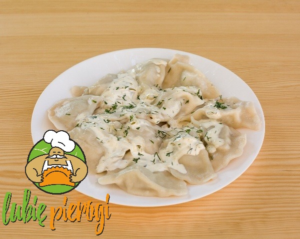 Porcja pierogów z sosem śmietanowym i koperkiem na białym talerzu, widoczne logo firmy 'lubie pierogi' w lewym dolnym rogu.