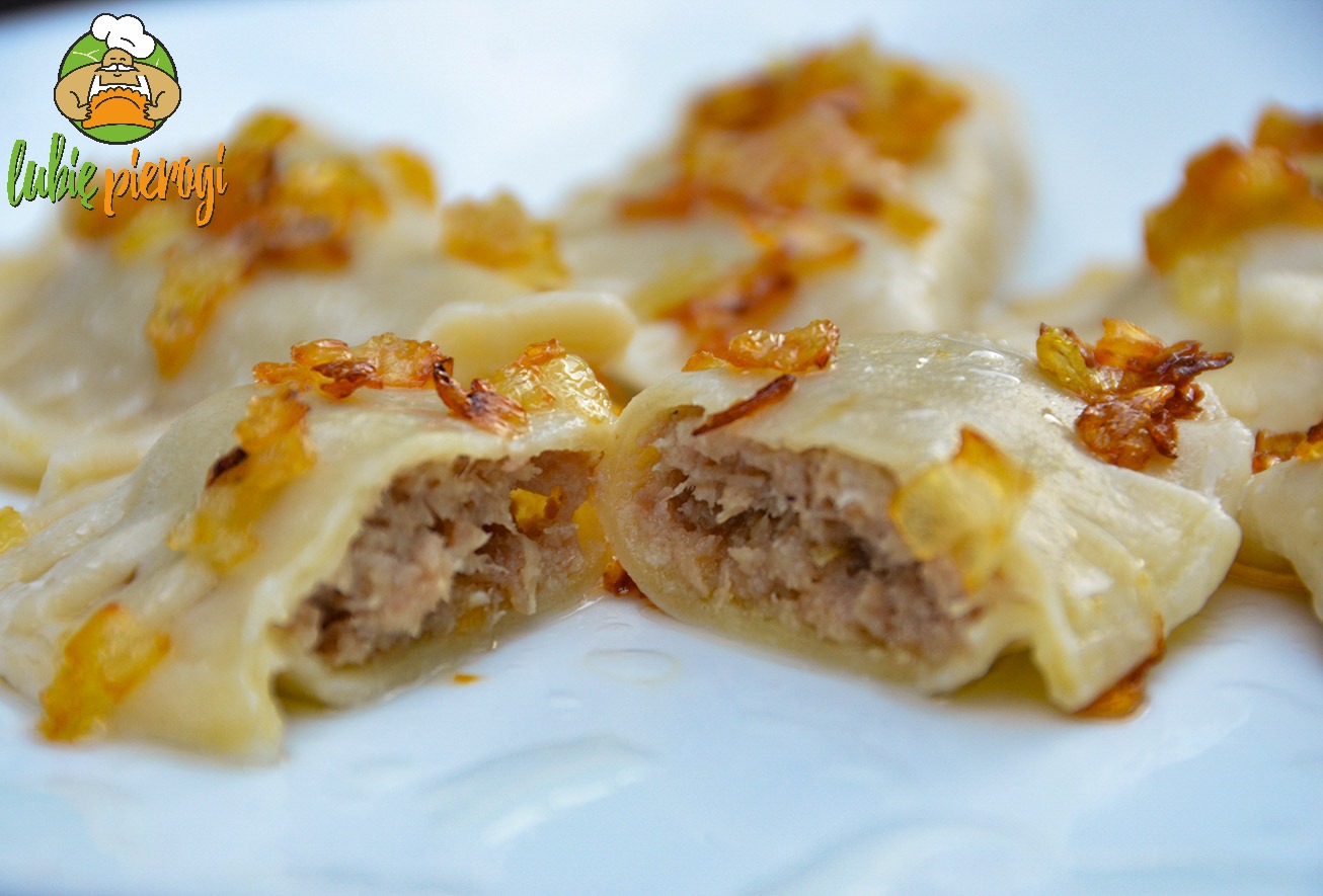 Zbliżenie na ugotowane pierogi z mięsem, posypane smażoną cebulką, jeden przekrojony na pół ukazujący farsz, ułożone na białym talerzu.