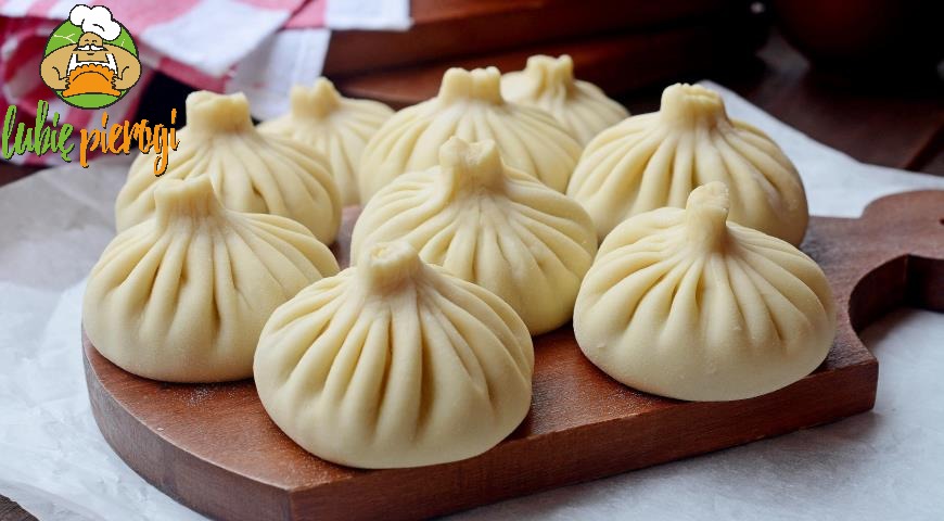 Dziewięć surowych chinkali ułożonych na drewnianej desce, z logo 'lubie pierogi' w lewym górnym rogu, na tle białej ściereczki i drewnianych elementów.