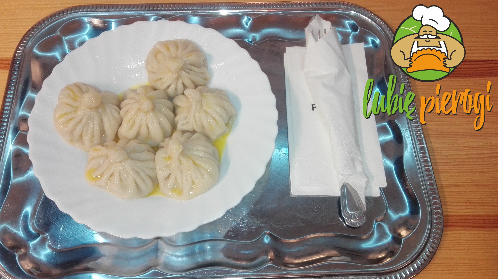 Sześć chinkali polanych masłem na białym, karbowanym talerzu, obok serwetki i sztućce na srebrnej tacy z dekoracyjnym brzegiem, w tle drewniany blat stołu i naklejka z logotypem.