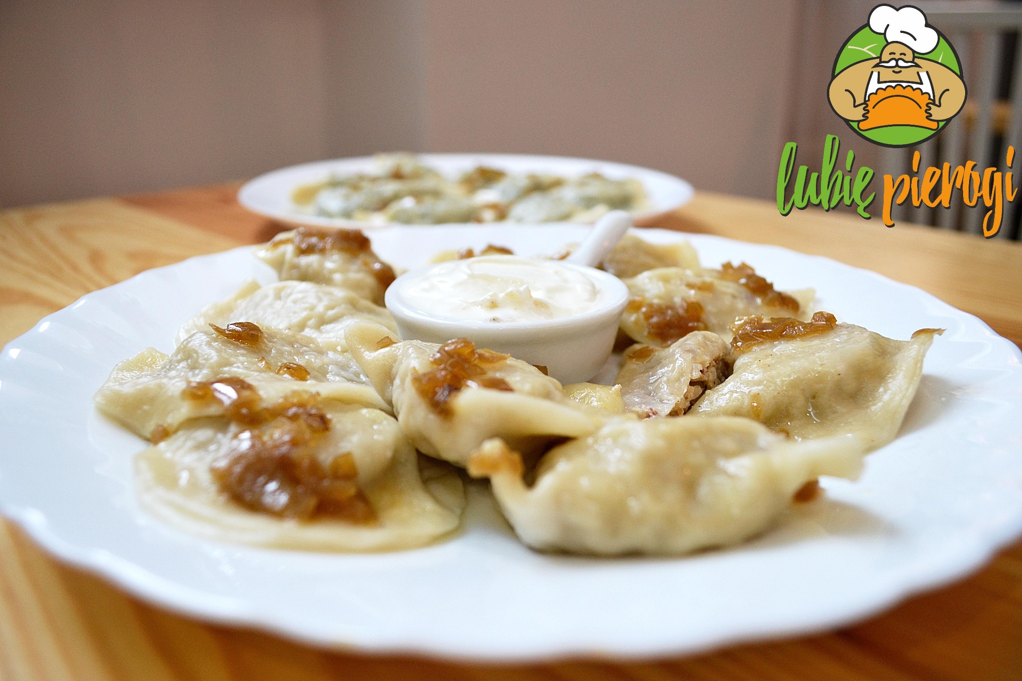 Pierogi z cebulką na białym talerzu, obok miseczka ze śmietaną, w tle drugi talerz z pierogami i logo firmy 'lubię pierogi'.