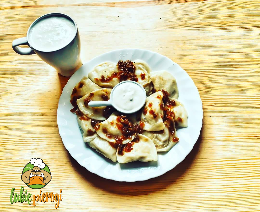 Pierogi okraszone cebulką na białym talerzu, obok kubek z sosem i logo firmy 'Lubię Pierogi' na drewnianym stole.
