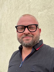 Portret mężczyzny w okularach, z krótką brodą, ubranego w czarną koszulkę polo z czerwonym wykończeniem, na tle jasnej ściany o chropowatej fakturze.