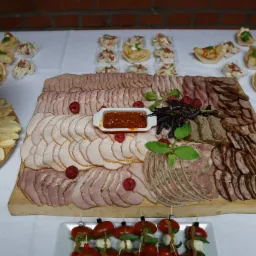 Bogato zastawiony stół z deską wędlin, serów, przekąsek na jeden kęs i sosem w białej miseczce, całość na białym obrusie na tle ściany z czerwonej cegły.