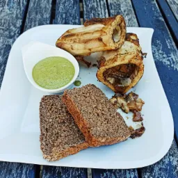 Pieczone szpik kostny z ciemnym pieczywem i zielonym sosem na białym kwadratowym talerzu, ułożone na niebieskim, drewnianym stole.