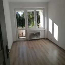 Pusty, świeżo odnowiony pokój z drewnianą podłogą, białymi ścianami i grzejnikiem pod oknem z widokiem na drzewa.