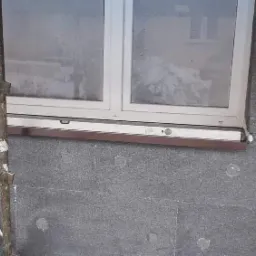 Szara elewacja budynku w trakcie ocieplania styropianem, widoczne okno z brązowym parapetem i fragment rusztowania