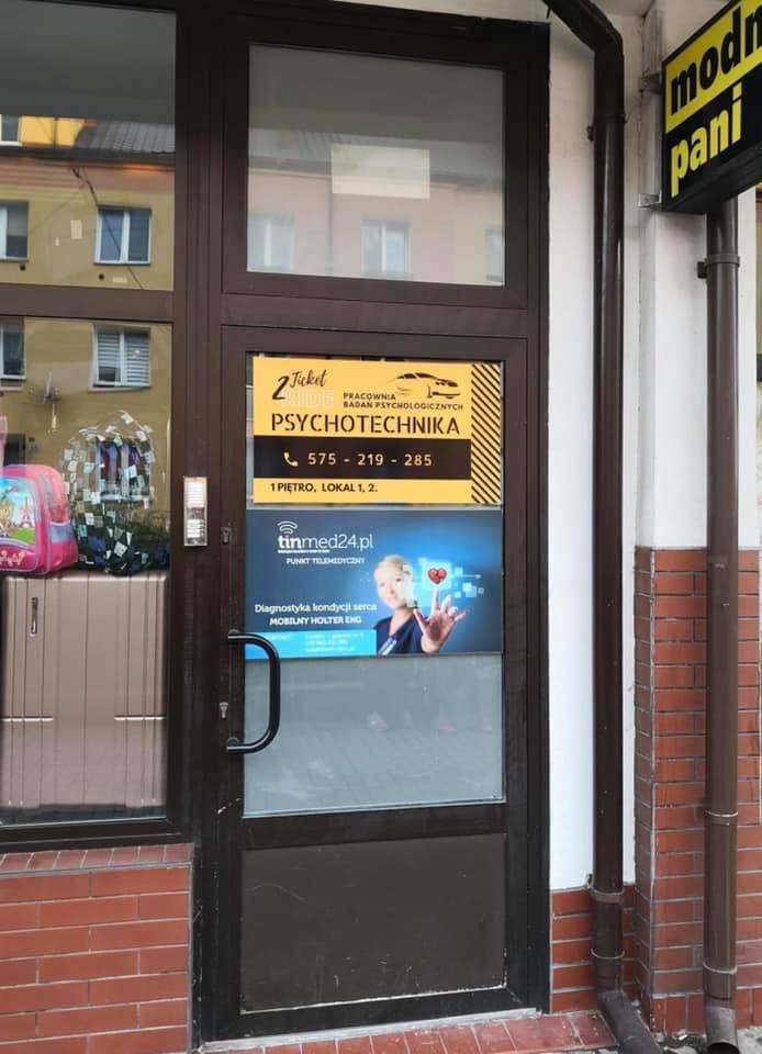 Wejście do pracowni badań psychologicznych i psychotechniki, na drzwiach żółta tabliczka z nazwą firmy i numerem telefonu, poniżej reklama punktu telemedycznego z kobietą dotykającą wirtualnego serca.