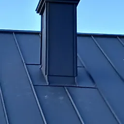 Szczegółowe ujęcie ciemnej, metalowej obróbki komina na dachu z blachy na rąbek stojący, widok z dołu pod kątem na tle błękitnego nieba.