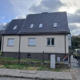 Dom jednorodzinny z ciemną dachówką i trzema oknami dachowymi, widok z ulicy, ogrodzenie z metaloplastyki.