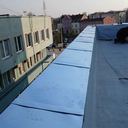 Uslugi dekarskie blacharskie Maciej Niewczas - Błyszczące, metalowe panele na płaskim dachu budynku, częściowo pokryte, z widocznym pracownikiem w prawym rogu i budynkami w tle. Widoczne ślady stóp na panelach.