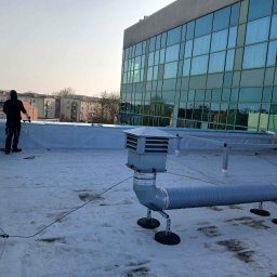Uslugi dekarskie blacharskie Maciej Niewczas - Praca na dachu płaskim: dekarz przy montażu instalacji wentylacyjnej. Widoczne narzędzia i budynek biurowy w tle. Jasne niebo.