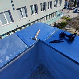 Uslugi dekarskie blacharskie Maciej Niewczas - Szary dach budynku po obróbkach blacharskich. Na dachu leżą narzędzia: młotek, nożyce do blachy i wkrętarka. W tle budynek z oknami i drzewa.