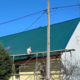 Uslugi dekarskie blacharskie Maciej Niewczas - Praca na dachu z zielonej blachy falistej. Dekarz na rusztowaniu przy kominie, obok słup elektryczny. Widok na dom z drewnianym płotem.