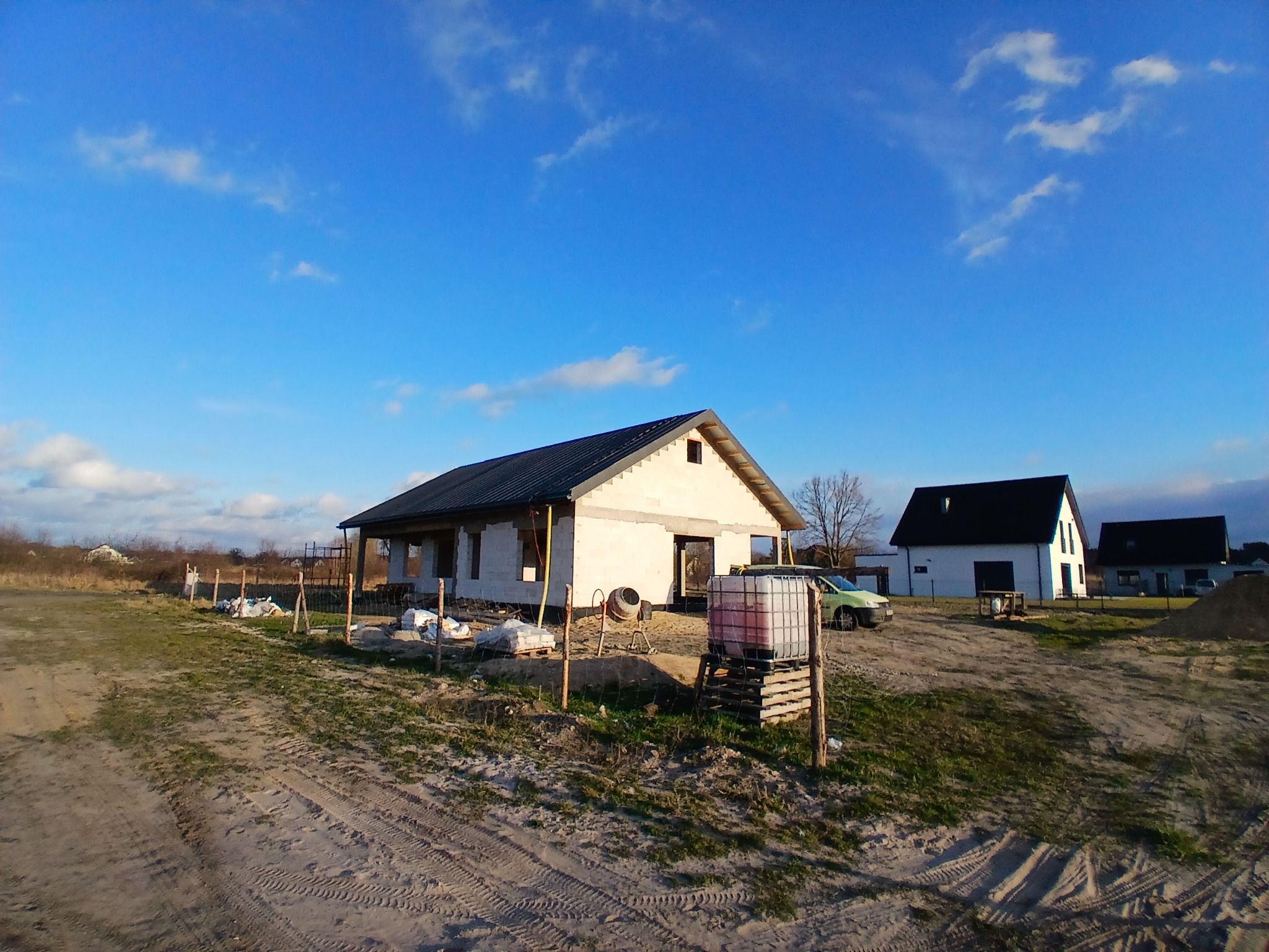 Surowy dom jednorodzinny z ciemnym dachem w trakcie budowy, widoczne rusztowania, materiały budowlane i zbiornik na wodę, na pierwszym planie piaszczysta ziemia.