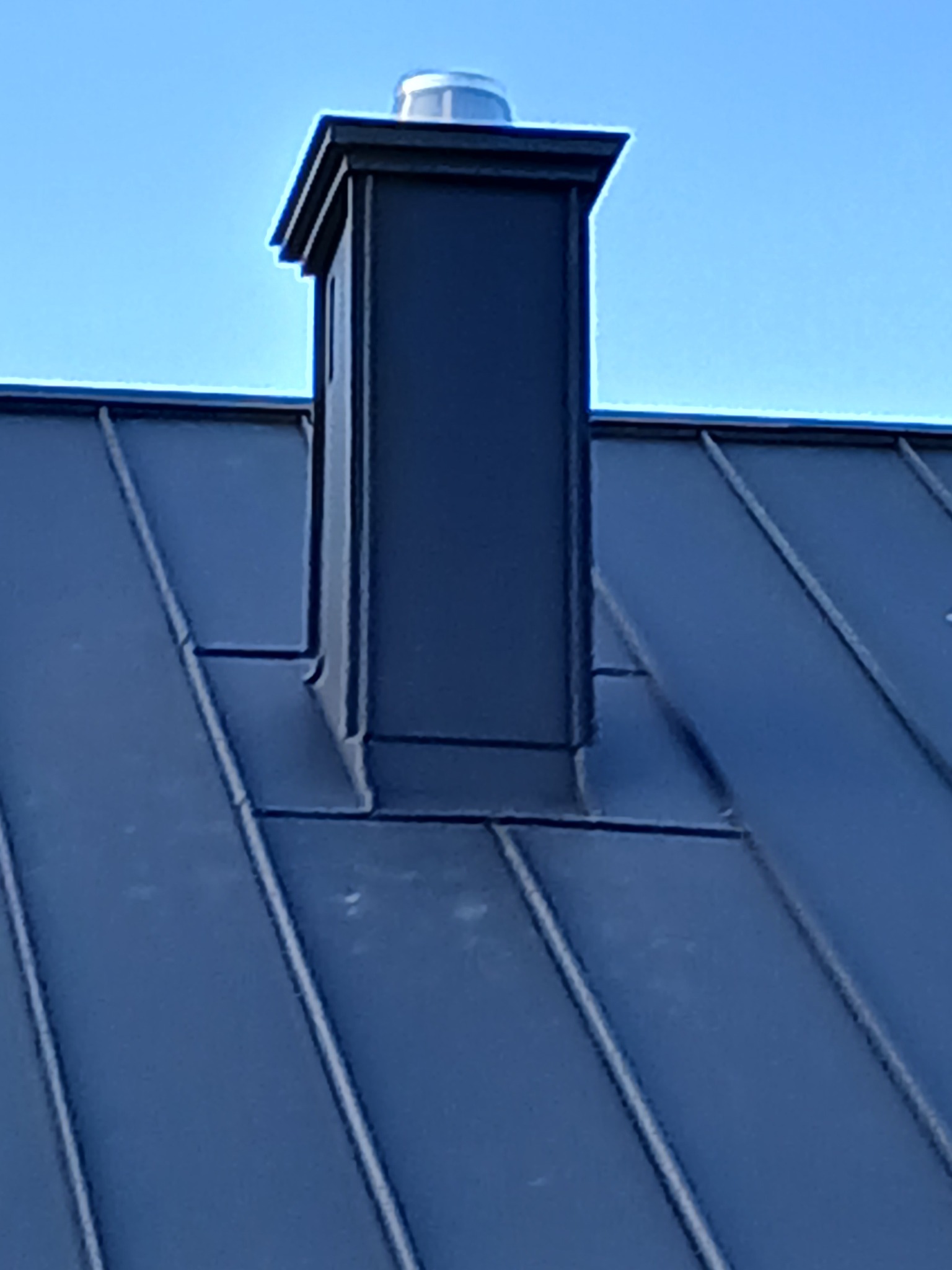 Szczegółowe ujęcie ciemnej, metalowej obróbki komina na dachu z blachy na rąbek stojący, widok z dołu pod kątem na tle błękitnego nieba.