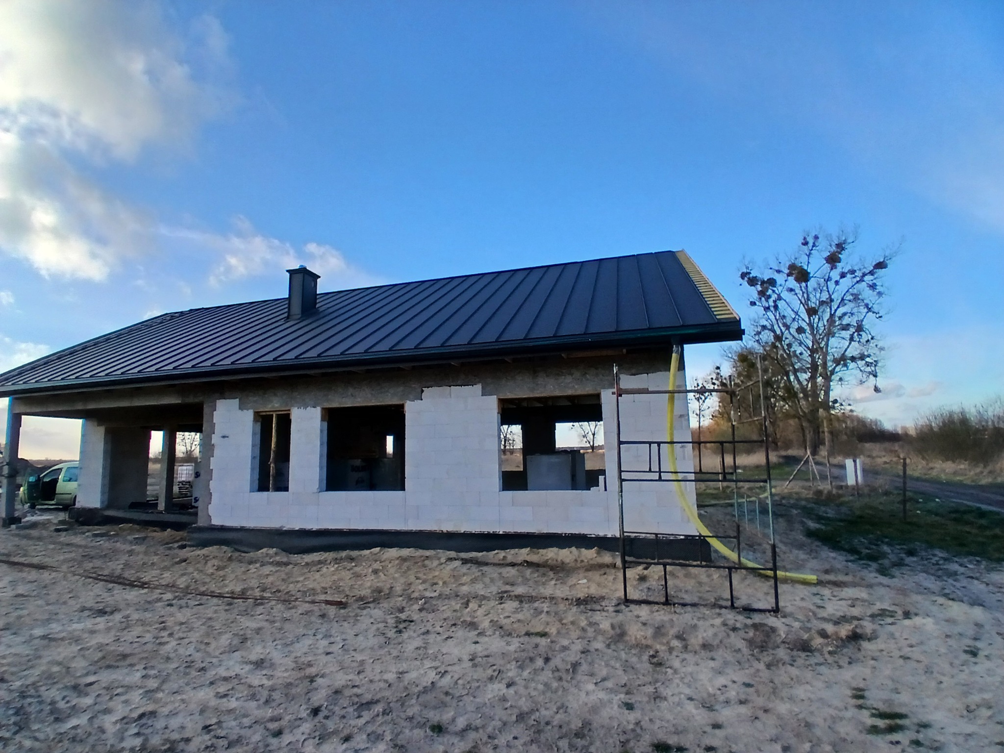 Dom w trakcie budowy z zamontowanym, ciemnym dachem z blachy na tle błękitnego nieba. Widoczne rusztowanie i materiały budowlane.