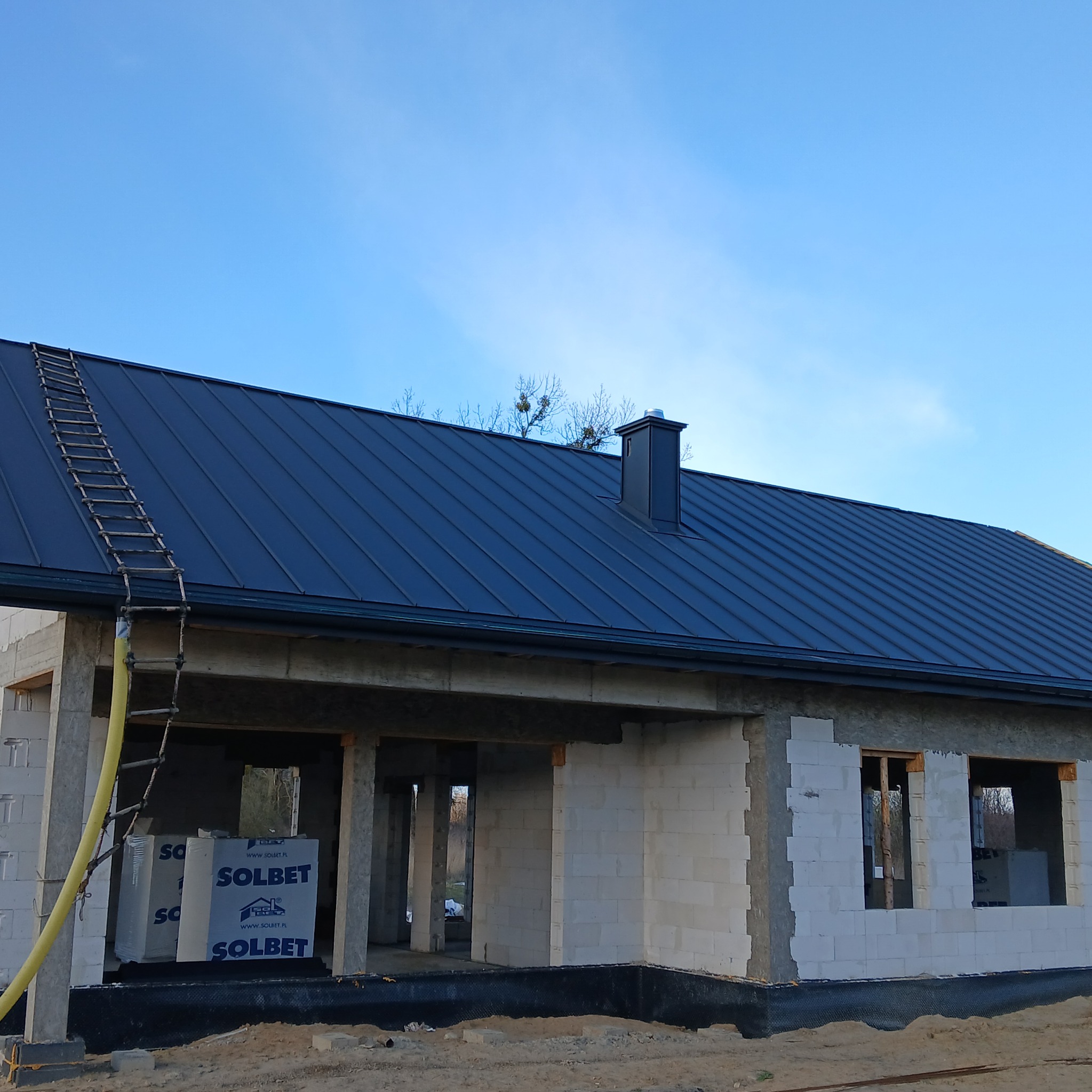 Nowy, ciemny dach z blachy na surowym budynku w trakcie budowy, drabina oparta o rynnę, pakiety bloczków SOLBET obok ściany.