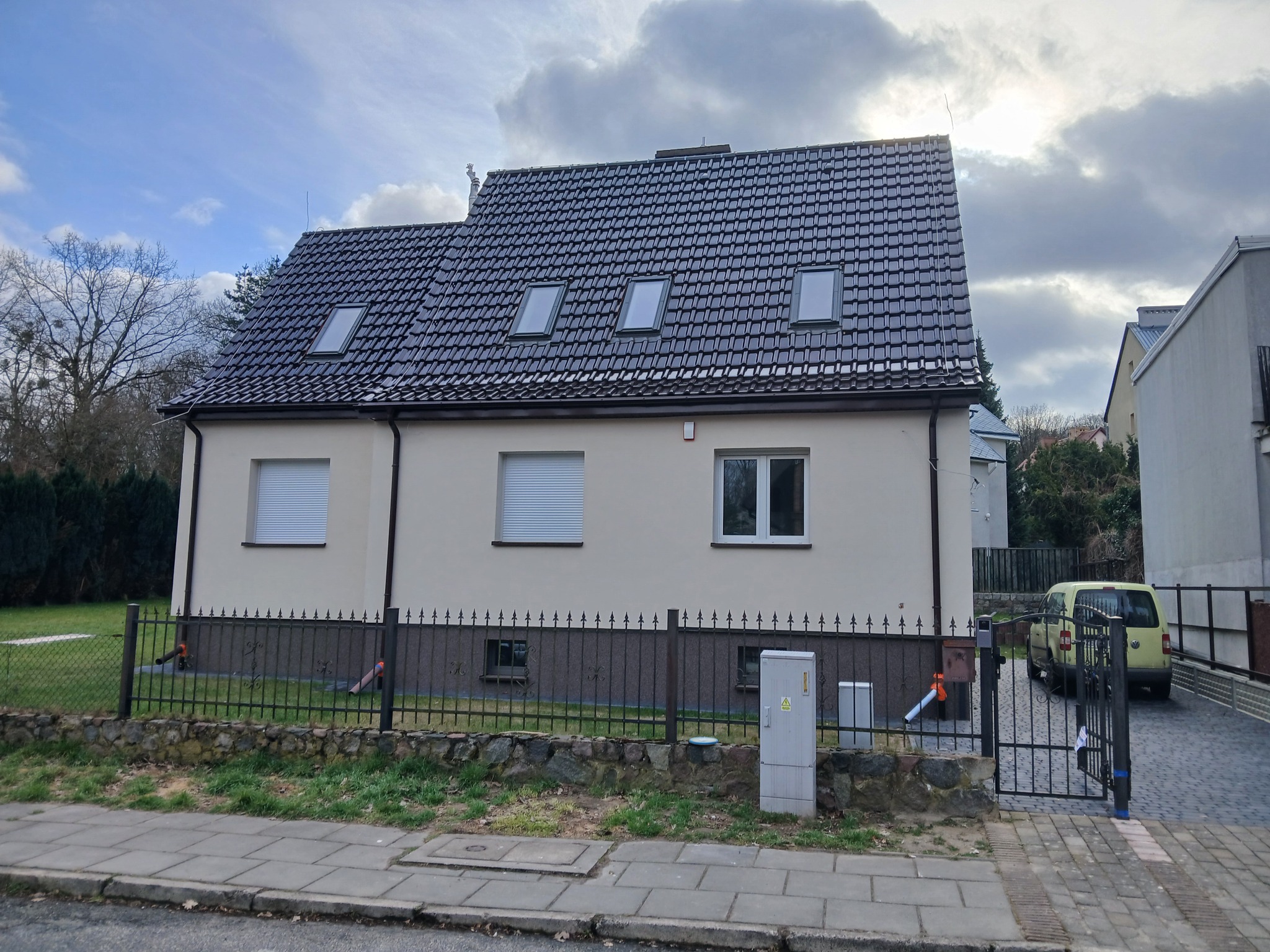 Dom jednorodzinny z ciemną dachówką i trzema oknami dachowymi, widok z ulicy, ogrodzenie z metaloplastyki.