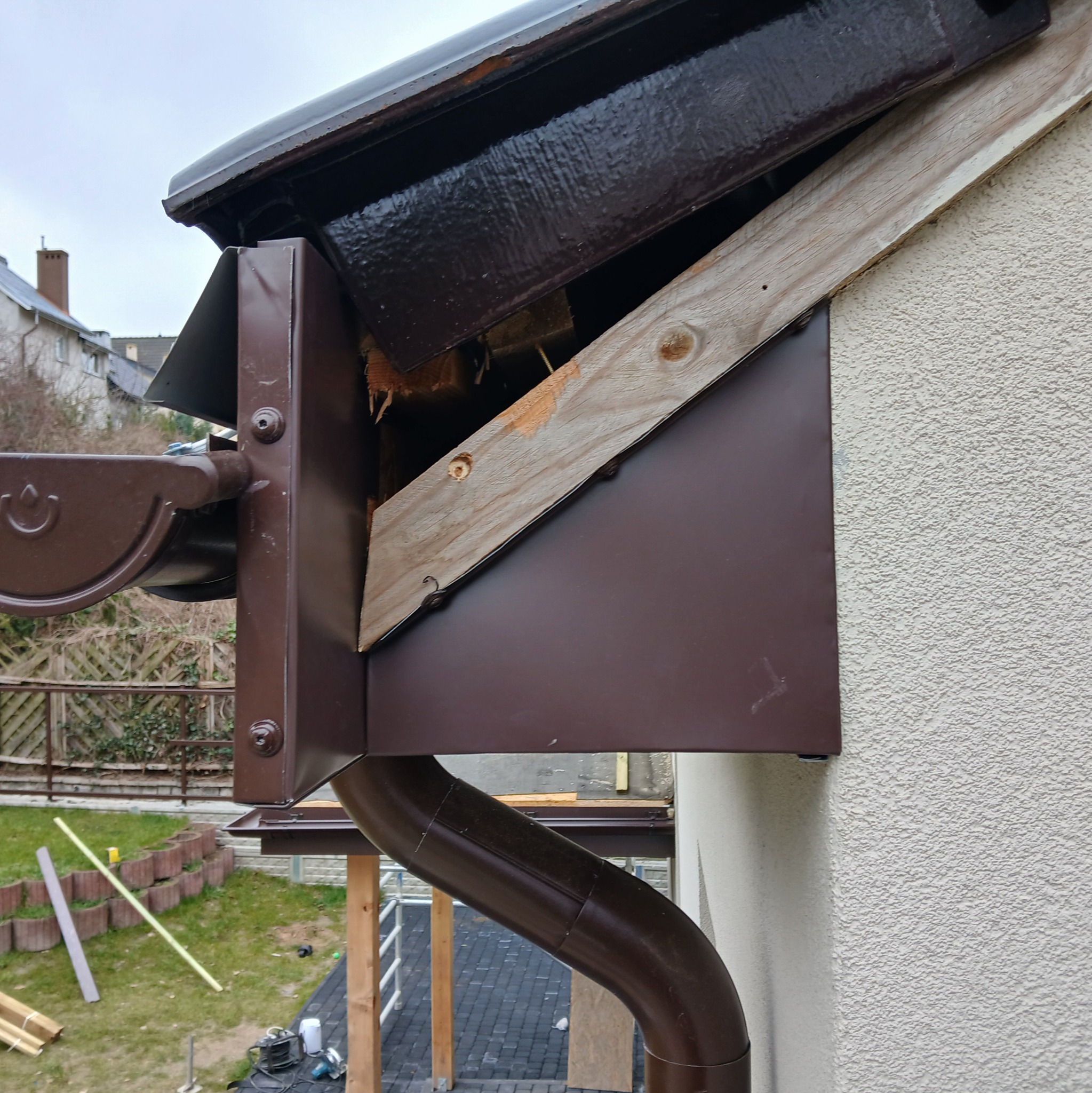 Brązowa rynna spustowa z elementami mocującymi, widoczne drewniane elementy konstrukcyjne, elewacja budynku z fakturą baranka.
