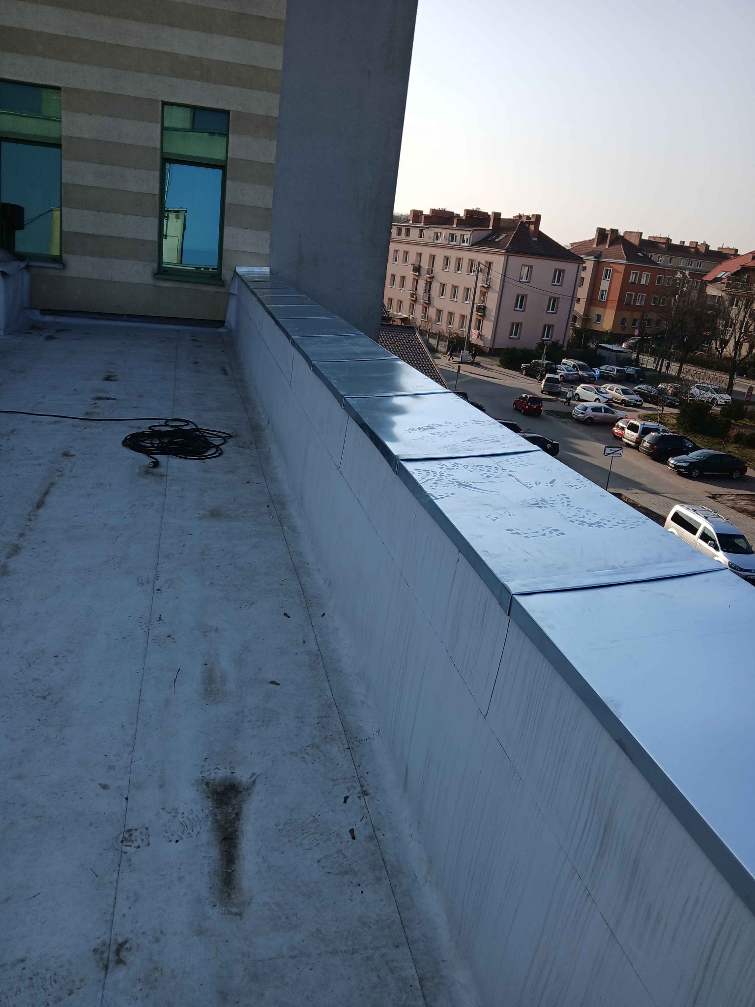 Wykończona obróbka blacharska na dachu budynku, widoczne ślady stóp na powierzchni. W tle zabudowa miejska i błękitne niebo.