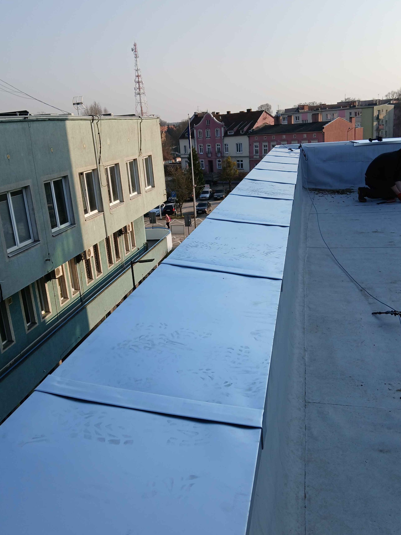 Błyszczące, metalowe panele na płaskim dachu budynku, częściowo pokryte, z widocznym pracownikiem w prawym rogu i budynkami w tle. Widoczne ślady stóp na panelach.