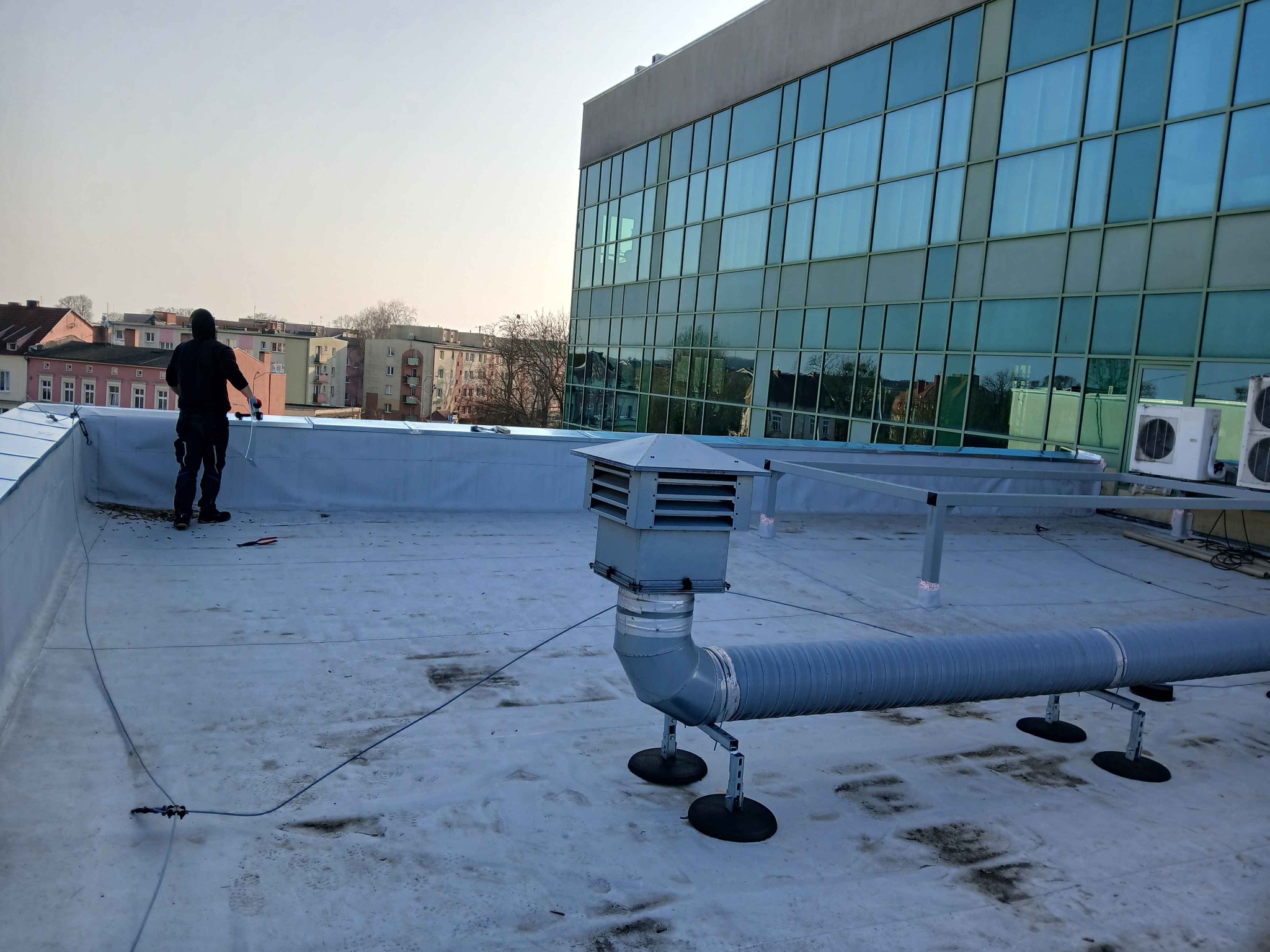 Praca na dachu płaskim: dekarz przy montażu instalacji wentylacyjnej. Widoczne narzędzia i budynek biurowy w tle. Jasne niebo.