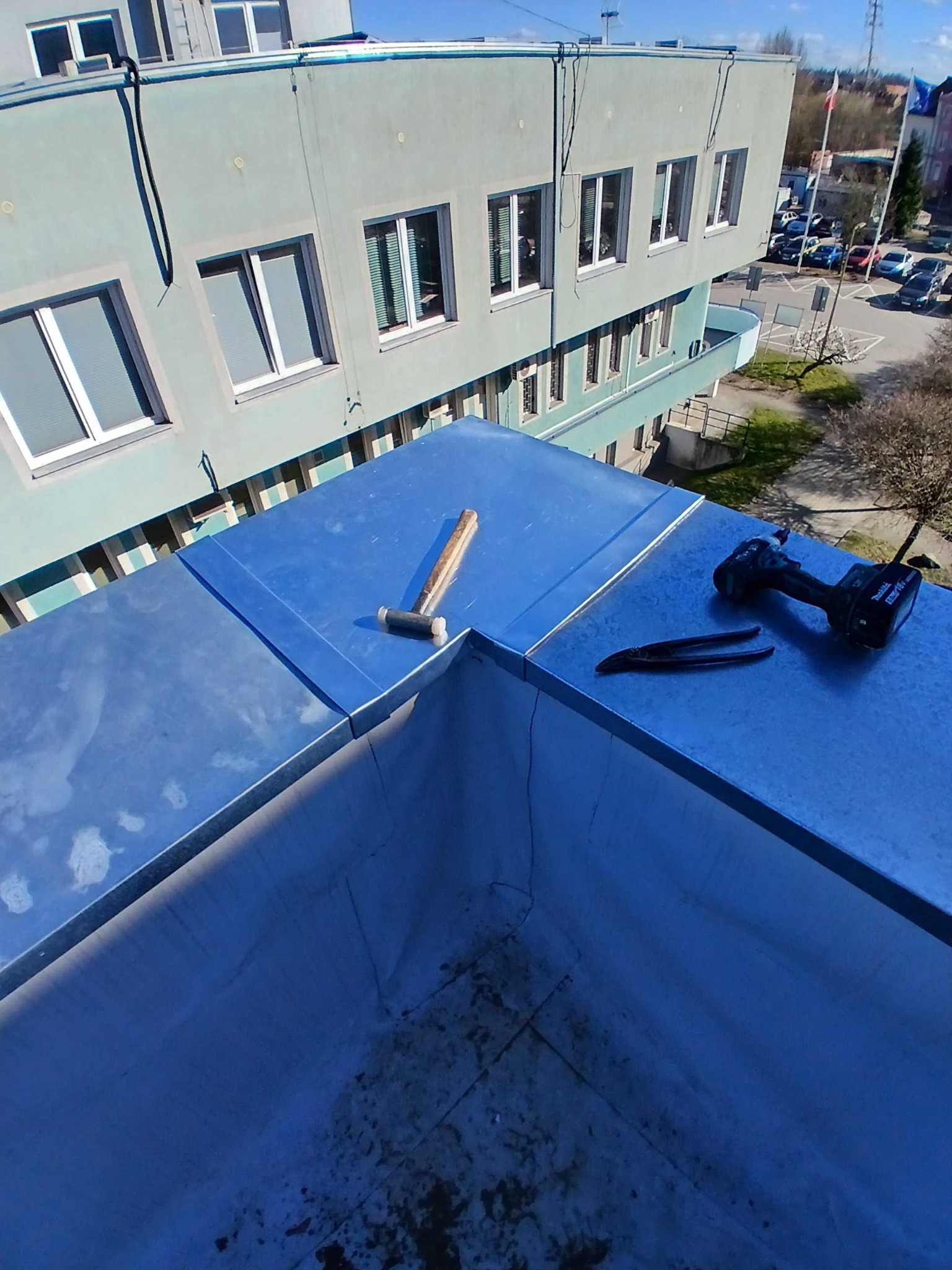 Szary dach budynku po obróbkach blacharskich. Na dachu leżą narzędzia: młotek, nożyce do blachy i wkrętarka. W tle budynek z oknami i drzewa.